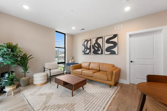 Inviting modern living room with elegant decor, warm leather sofa, abstract art, and large window offering cityscape views.