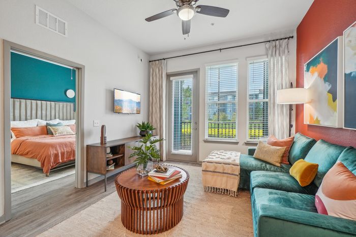 Inviting and stylish living room with vibrant artwork, plush teal sofa, and abundant natural light, adjoining a cozy bedroom with colorful bedding.
