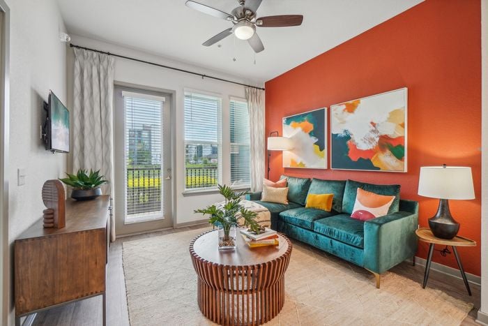 Vibrant living room with modern decor, featuring a teal velvet sofa, bold orange accent wall, and abstract art. Sunlit space with glass doors, curtain-draped windows, and a chic wooden coffee table.