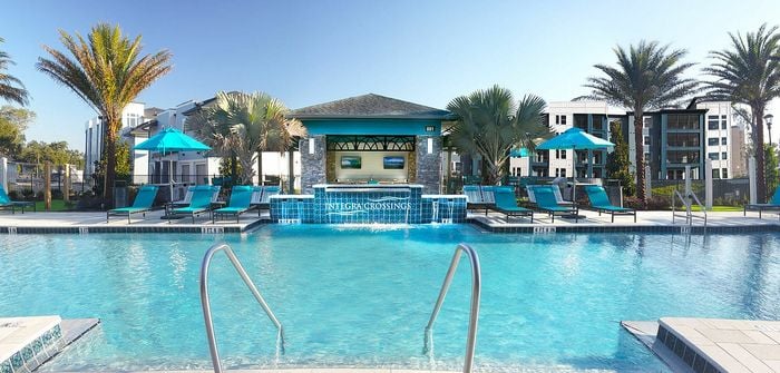 Luxurious apartment complex featuring a stunning pool area with palm trees, elegant lounge chairs, and vibrant blue umbrellas.