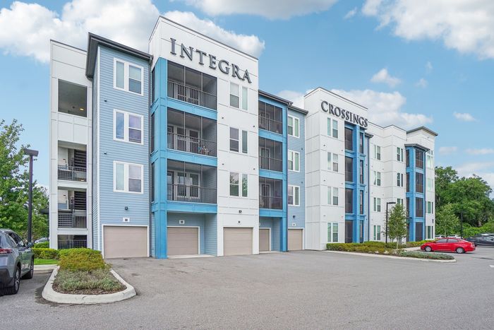Modern apartment complex featuring stylish multi-story buildings with blue accents, ample parking, and lush green landscaping under a bright sky.
