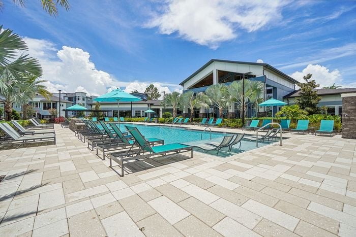 Luxurious apartment complex featuring a pristine outdoor pool area with comfortable lounge chairs and vibrant teal umbrellas under a clear blue sky.