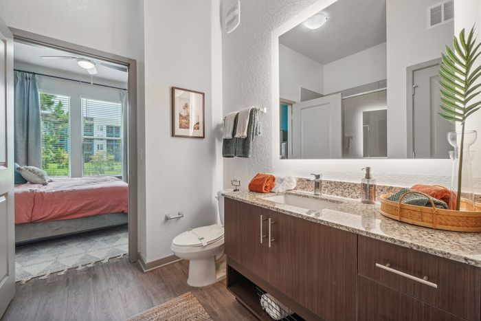 Modern bathroom with granite countertop, illuminated mirror, and view into cozy bedroom with large windows and pink bedding.