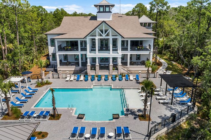 Luxurious two-story clubhouse surrounded by lush greenery, featuring a pristine pool with stylish blue lounge chairs and elegant outdoor seating.