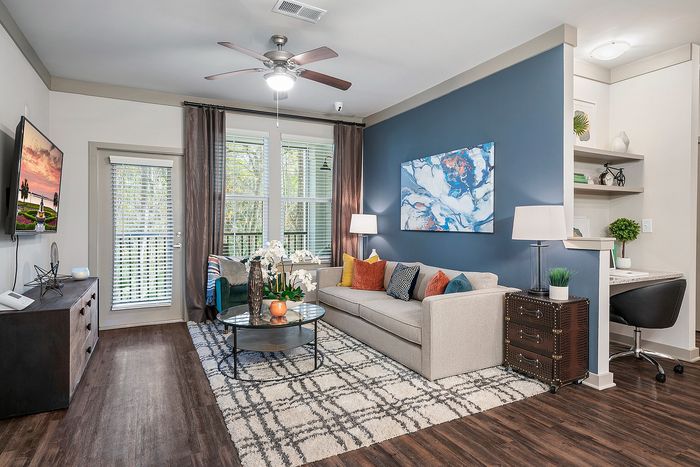 Elegant living room with a cozy beige sofa, vibrant cushions, and a chic marble-top coffee table on a stylish patterned rug. Features a striking blue accent wall with modern art and generous natural light through large windows.