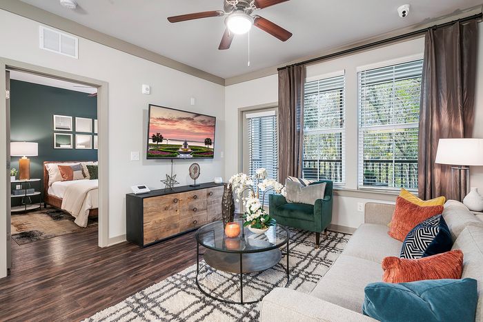 Inviting living space with elegant decor, featuring a cozy sofa, vibrant cushions, and a view into a stylish bedroom. Large windows offer abundant natural light, enhancing the modern, serene atmosphere.