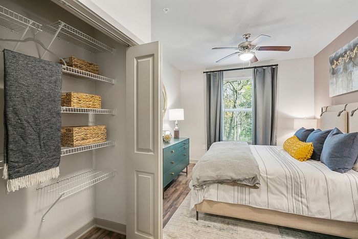 Bright and cozy bedroom with stylish decor, featuring a comfortable bed, modern dresser, and a spacious closet filled with woven storage baskets. The large window offers a serene view, enhancing the inviting atmosphere.