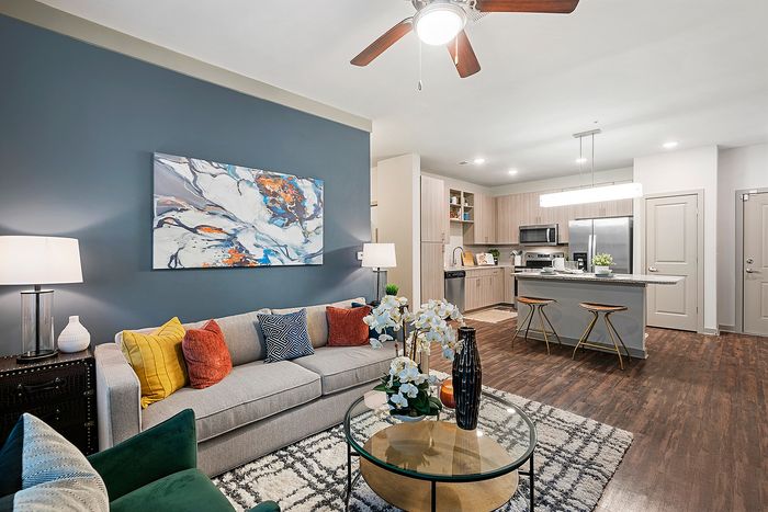 Modern living room with stylish decor, featuring a comfy gray sofa, vibrant accent pillows, chic wall art, and open kitchen with sleek cabinets.