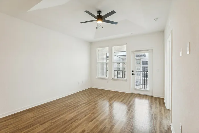 Bright and inviting room with elegant wooden flooring, large windows, and a modern ceiling fan, creating a spacious ambiance perfect for comfortable living.