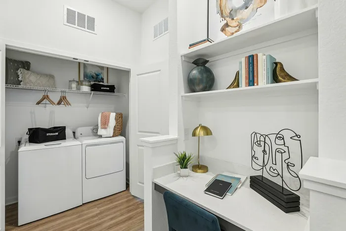 Charming apartment corner featuring a modern laundry closet with a washer and dryer alongside a chic workspace with decorative shelving and contemporary art pieces.