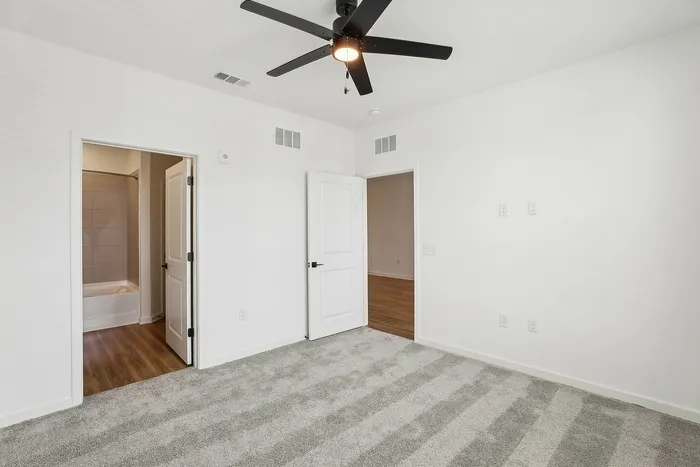 Bright and inviting bedroom with plush carpet, modern ceiling fan, and convenient access to a tiled bathroom. Ideal for comfortable living.