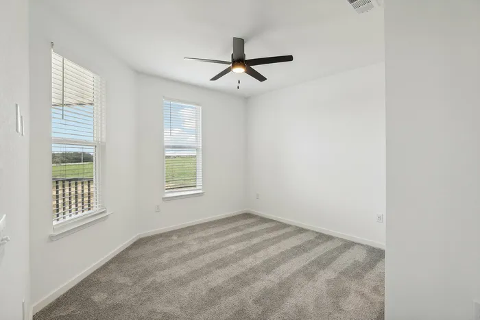 Bright and airy room with plush carpeting, large windows, and a modern ceiling fan, offering a serene view of open greenery.