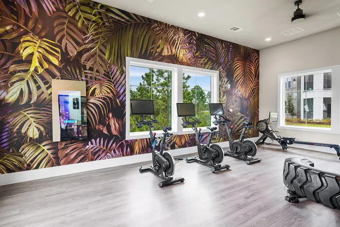 Modern fitness room with vibrant botanical wallpaper, featuring exercise bikes and rowing machines, brightened by natural light from large windows.