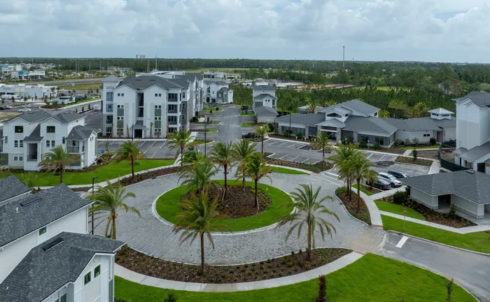 Aerial view of a modern residential complex with stylish buildings, well-maintained landscaping, palm trees, and spacious parking area under a cloudy sky.