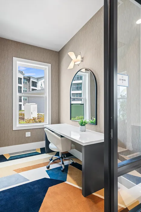 Modern coworking space with sleek white desk, plush geometric carpet, and natural light through large window.