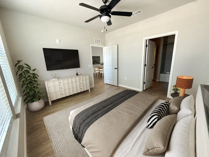 Bright and inviting bedroom with modern decor featuring a sleek ceiling fan, wall-mounted TV, and cozy bed adorned with stylish cushions. Natural light pours in through large windows, offering a serene ambiance.