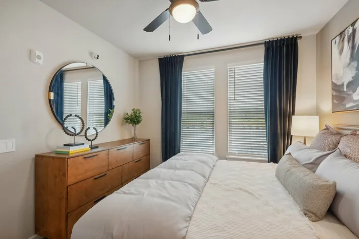 Elegant bedroom with a plush bed, stylish wooden dresser topped with decor, and large windows adorned with chic dark curtains, creating a serene and inviting ambiance.