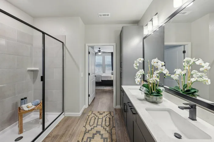 Elegant modern bathroom with a sleek glass shower, stylish double vanity, and orchid decor, leading to a cozy bedroom.