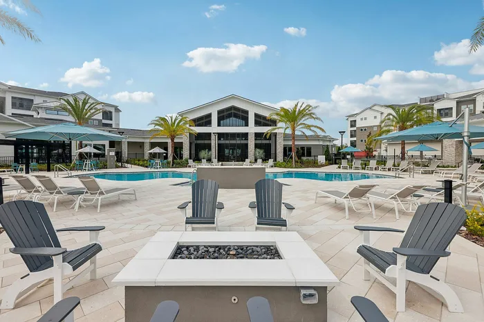 Luxurious apartment complex with inviting pool, modern lounge chairs, and elegant landscaping under a bright blue sky.