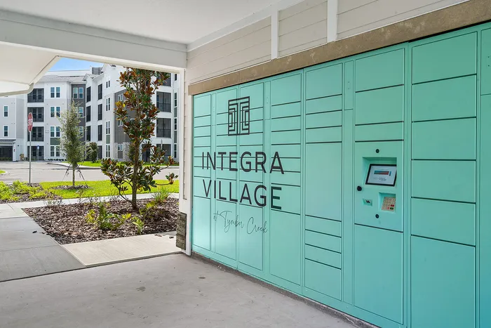 Contemporary entrance of Integra Village at Tymber Creek with vibrant turquoise parcel lockers, lush landscaping, and modern apartment buildings in the background.