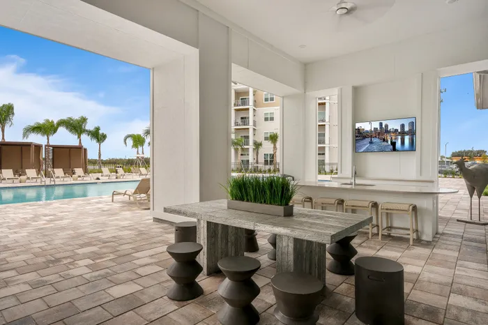 Luxurious outdoor oasis featuring a sleek dining area, modern bar with high chairs, and a sparkling pool surrounded by palm trees under a clear blue sky.
