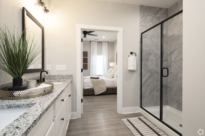 Elegant bathroom featuring a sleek shower, dual-sink granite vanity, and view into a cozy, well-lit bedroom with plush bedding.