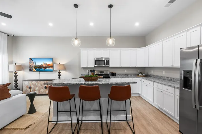 Modern open kitchen with sleek white cabinetry, granite countertops, and stylish pendant lighting. Features a breakfast bar with elegant brown stools and cozy seating area, ideal for entertaining.