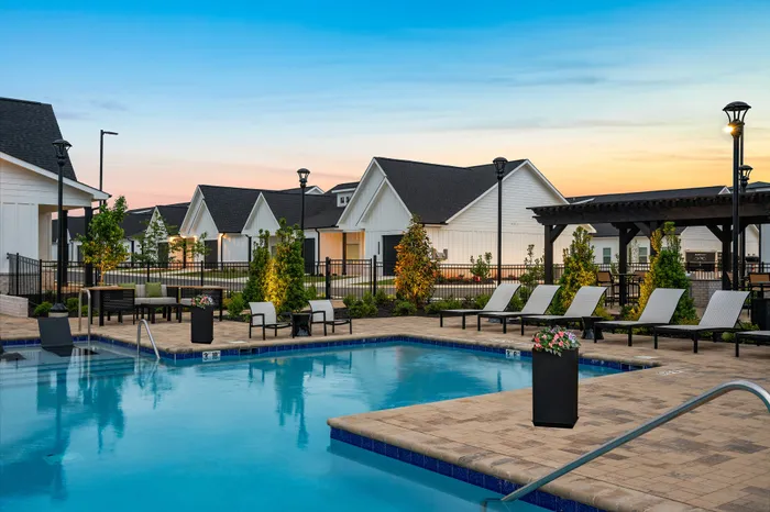 Elegant community pool area at sunset with modern lounge chairs, well-maintained landscaping, and charming white cottages in the backdrop.