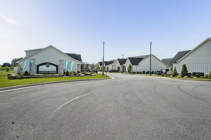 Charming villa community entrance with modern homes, manicured lawns, and welcoming signage on a sunny day.