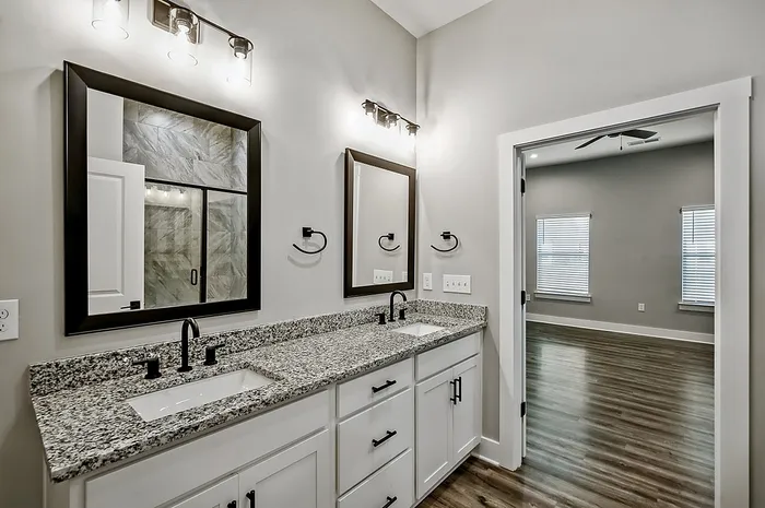 Sleek modern bathroom with dual granite vanity and stylish mirrors, leading to a bright spacious room with elegant wood flooring.