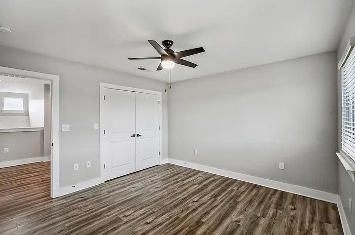 Bright and airy bedroom with modern ceiling fan and elegant wood floors, featuring a spacious closet and ample natural light streaming through the window.