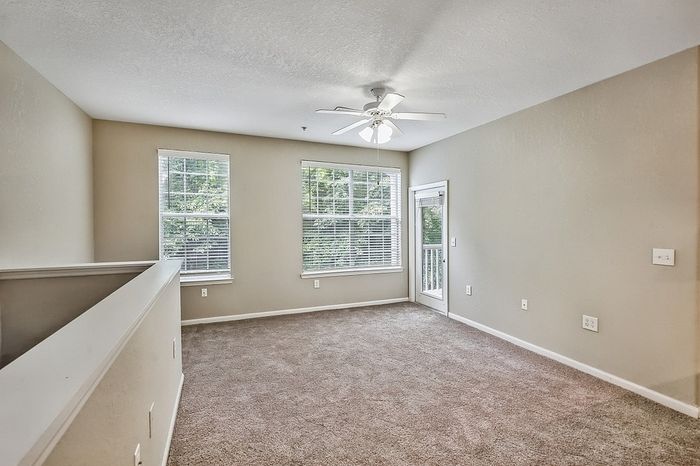 Bright and airy living room with plush carpeting, large windows, and ceiling fan, offering a serene view of greenery.