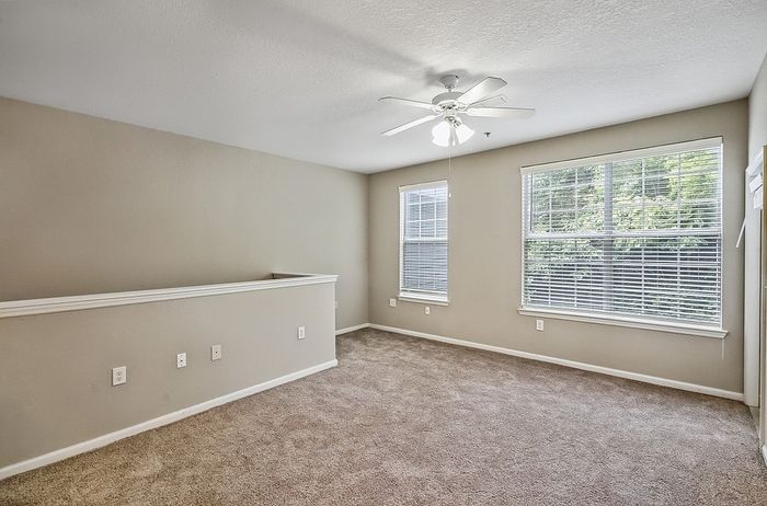 Inviting, sunlit apartment room with plush carpeting, ceiling fan, and large windows providing a serene view of lush greenery.