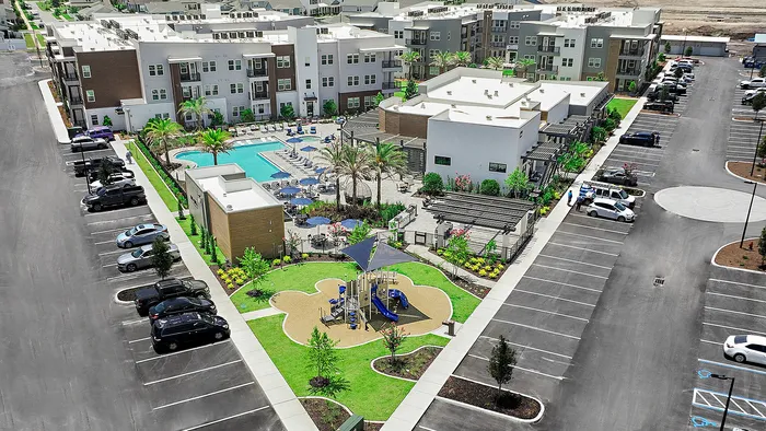 Aerial view of a modern apartment complex with lush landscaping, inviting pool area, and a vibrant children's playground, surrounded by ample parking space.