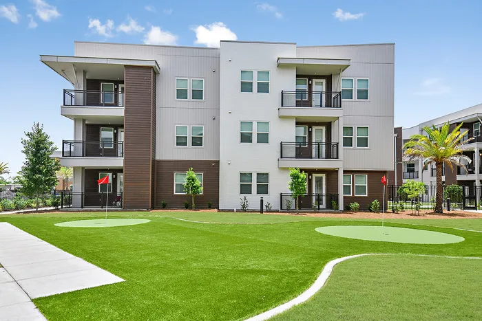 Elegant three-story apartment building featuring modern balconies and lush landscaping with a nearby putting green, set against a clear blue sky.