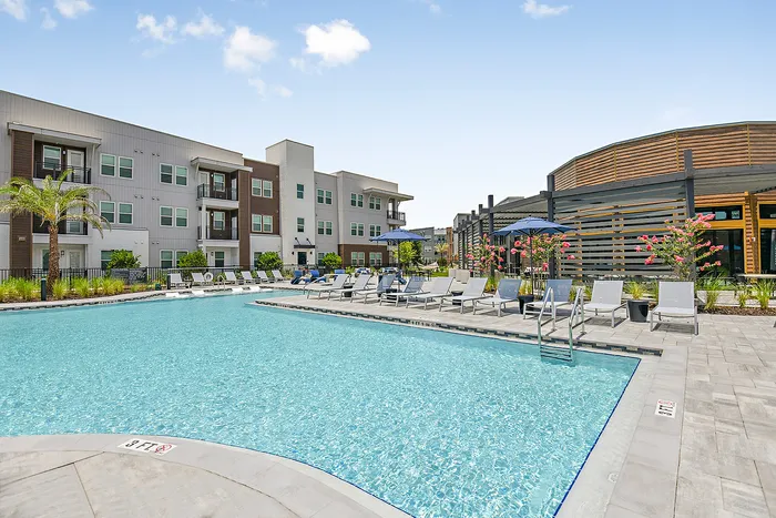 Luxurious apartment complex featuring a sparkling swimming pool, modern architecture, and relaxing lounge chairs under a clear blue sky.