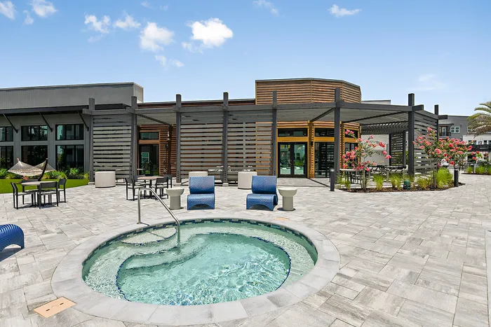 Luxurious apartment patio featuring a sparkling hot tub, modern outdoor seating, and lush landscaping under a clear blue sky.