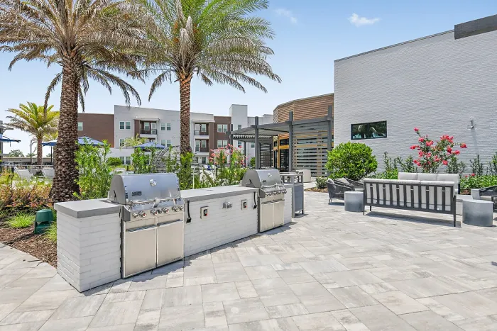 Modern outdoor patio area featuring sleek stainless-steel grills, lush palm trees, and stylish seating with vibrant greenery, perfect for social gatherings.
