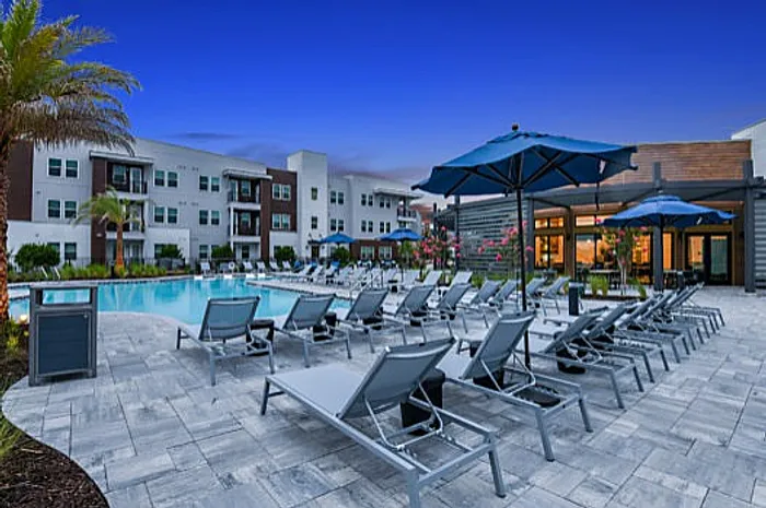 Luxurious apartment complex pool area with stylish lounge chairs and vibrant umbrellas, framed by modern buildings and lush landscaping under a twilight sky.