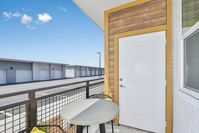 Charming apartment balcony with modern railing and cozy seating, offering a serene view of a row of sleek garage doors under a bright blue sky.