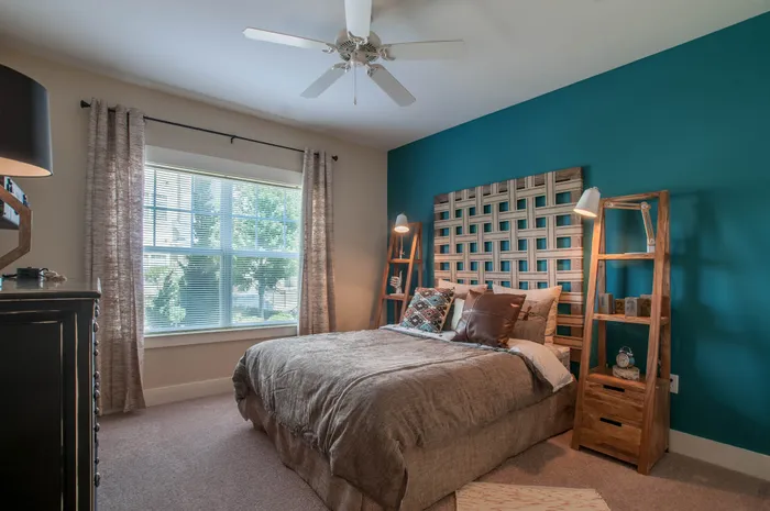 Cozy bedroom with a stylish wooden headboard, teal accent wall, and natural light pouring through large windows, creating a serene atmosphere.