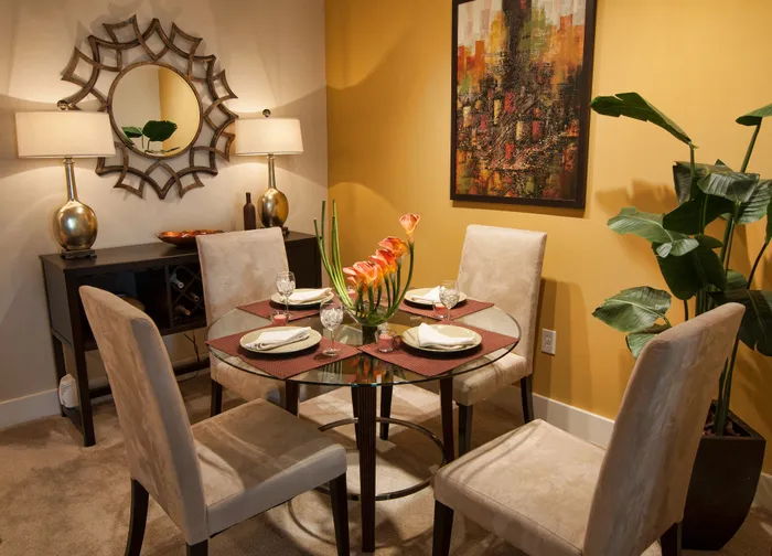 Elegant dining room with a round glass table set for four, featuring plush chairs, vibrant artwork, and stylish decor in a warm, inviting atmosphere.