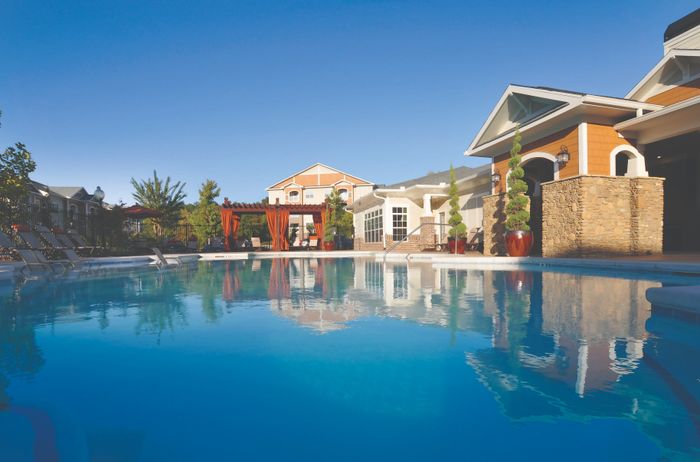 Luxurious apartment complex featuring a serene swimming pool, elegant pergola with red drapes, and beautifully landscaped surroundings on a sunny day.