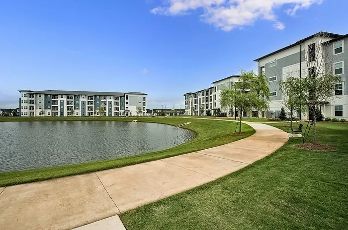 Charming apartment complex with modern architecture, overlooking a peaceful pond and surrounded by lush greenery on a sunny day.