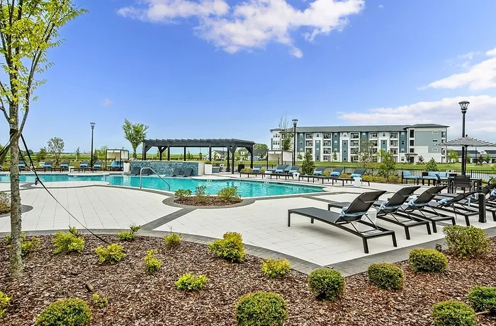 Luxurious apartment complex with a spacious outdoor pool area, surrounded by modern lounge chairs and lush landscaping, under a clear blue sky.