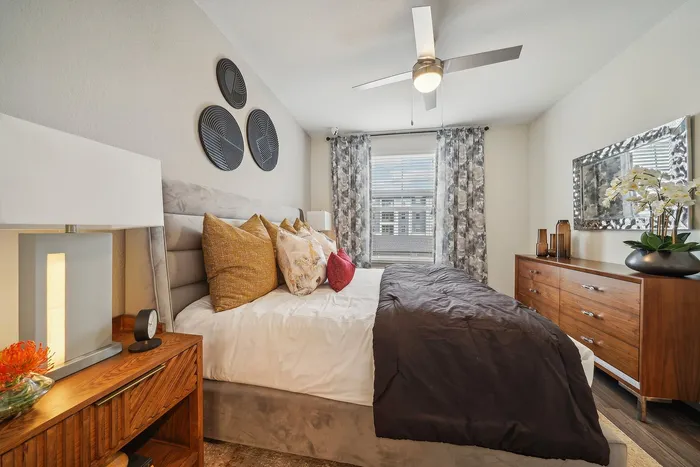 Elegant bedroom with modern decor, featuring a plush bed, chic cushions, stylish mirror, and sleek wooden furniture bathed in natural light.