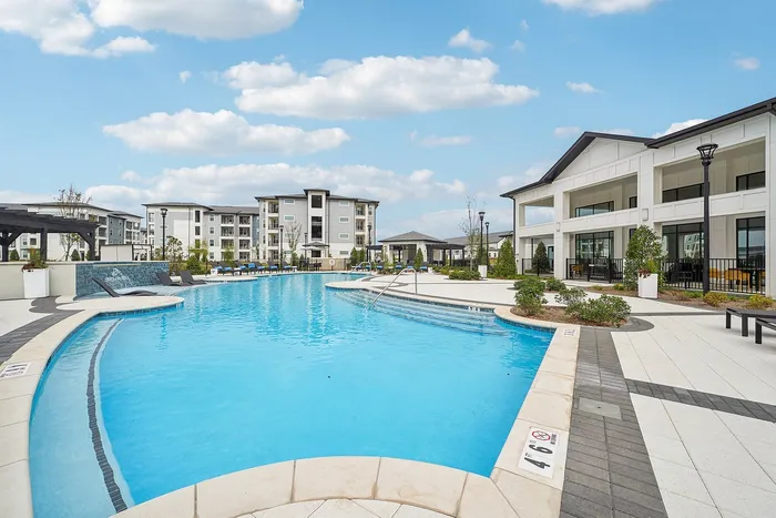 Luxurious apartment complex with a stunning blue pool, surrounded by stylish modern buildings and lush landscaping under a bright sky.