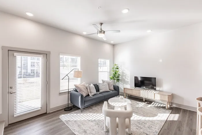 Bright and airy living room with modern furnishings, featuring a comfy gray sofa, chic floor lamp, sleek flat-screen TV, and abundant natural light through large windows.