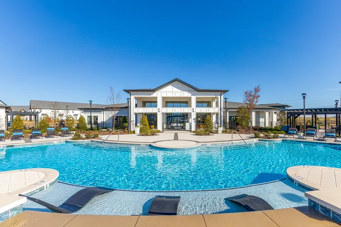 Luxurious apartment complex showcasing a stunning modern clubhouse with a refreshing swimming pool and inviting sun loungers set against a clear blue sky.