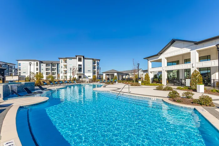 Luxurious apartment complex surrounded by a sparkling swimming pool, stylish lounge chairs, and modern architecture under a clear blue sky.